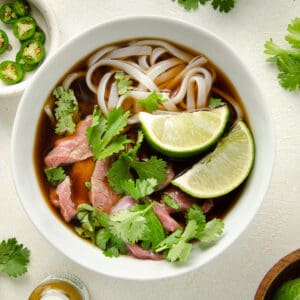 A bowl of gluten free pho with beef topped with two limes and cilantro.