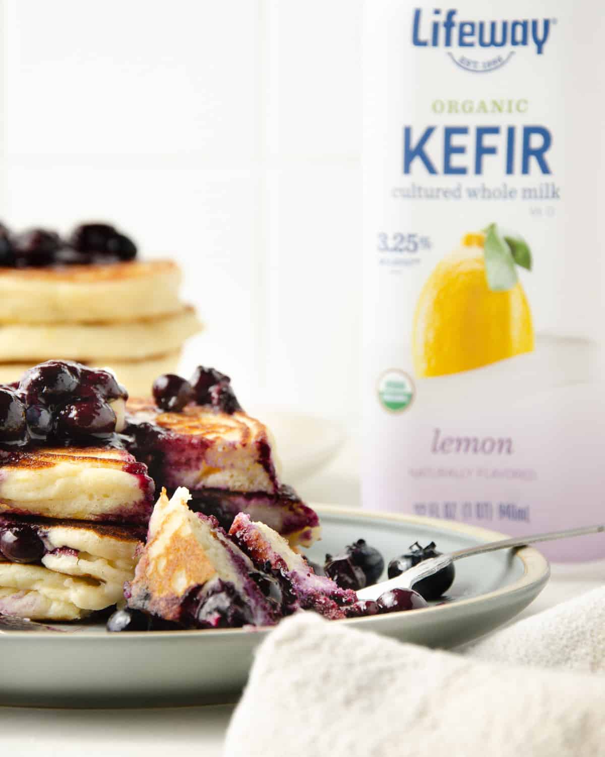 A plate of stacked blueberry kefir pancakes with a lemon kefir milk bottle to the side.