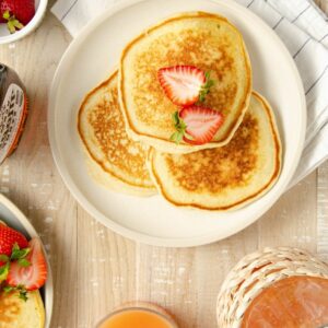 a beige plate of three pancakes topped with halved strawberry with a bowl of strawberries to the bottom, a second plate to the top left, and grapefruit juice