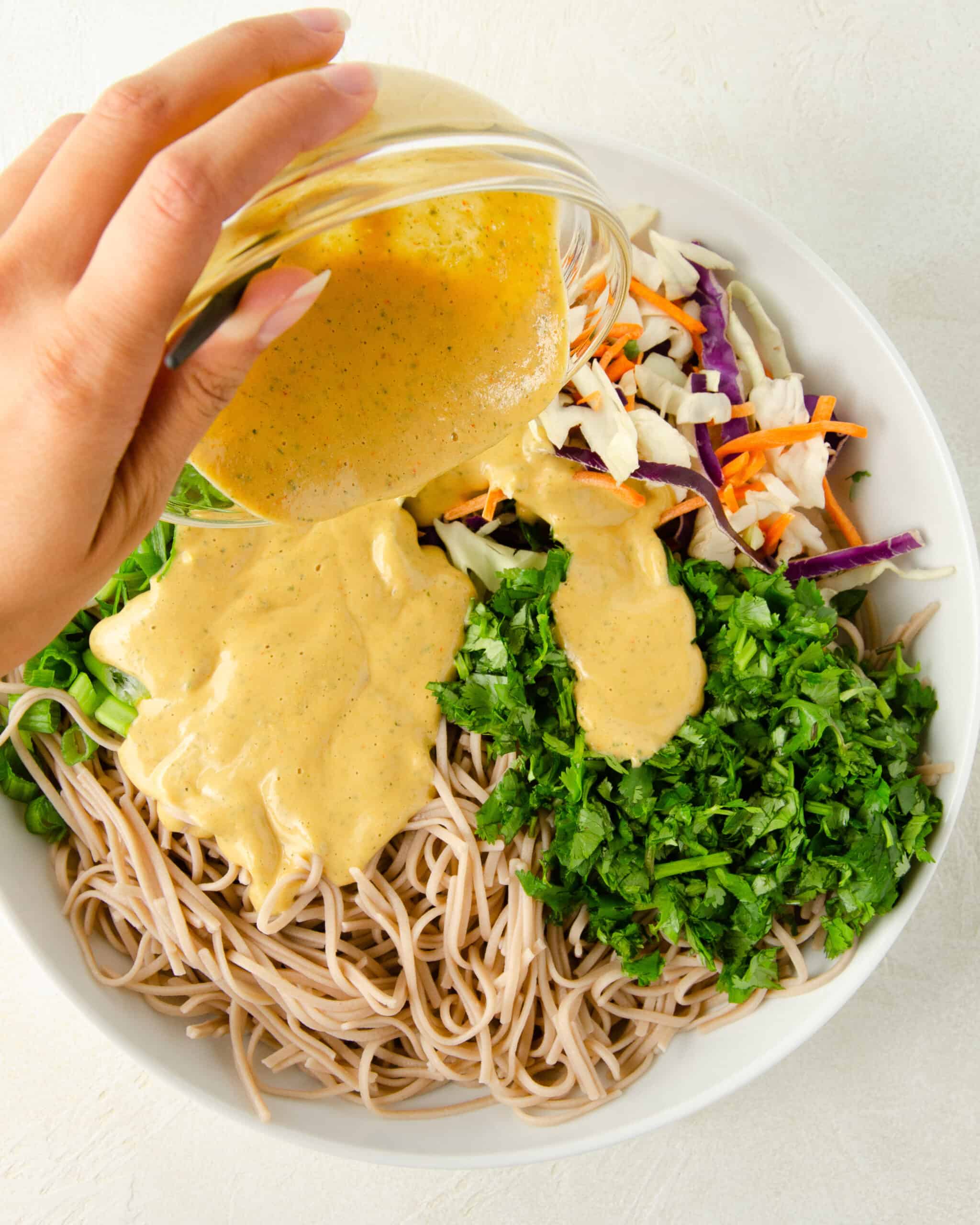 a hand pouring spicy cashew dressing into cold soba noodle salad