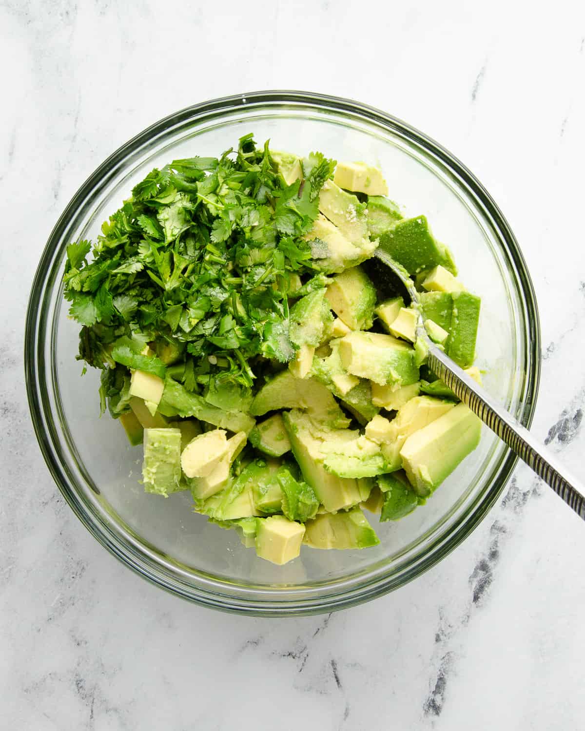Diced avocado, cilantro, garlic salt, and lime juice in a glass bowl on a white counter top. 
