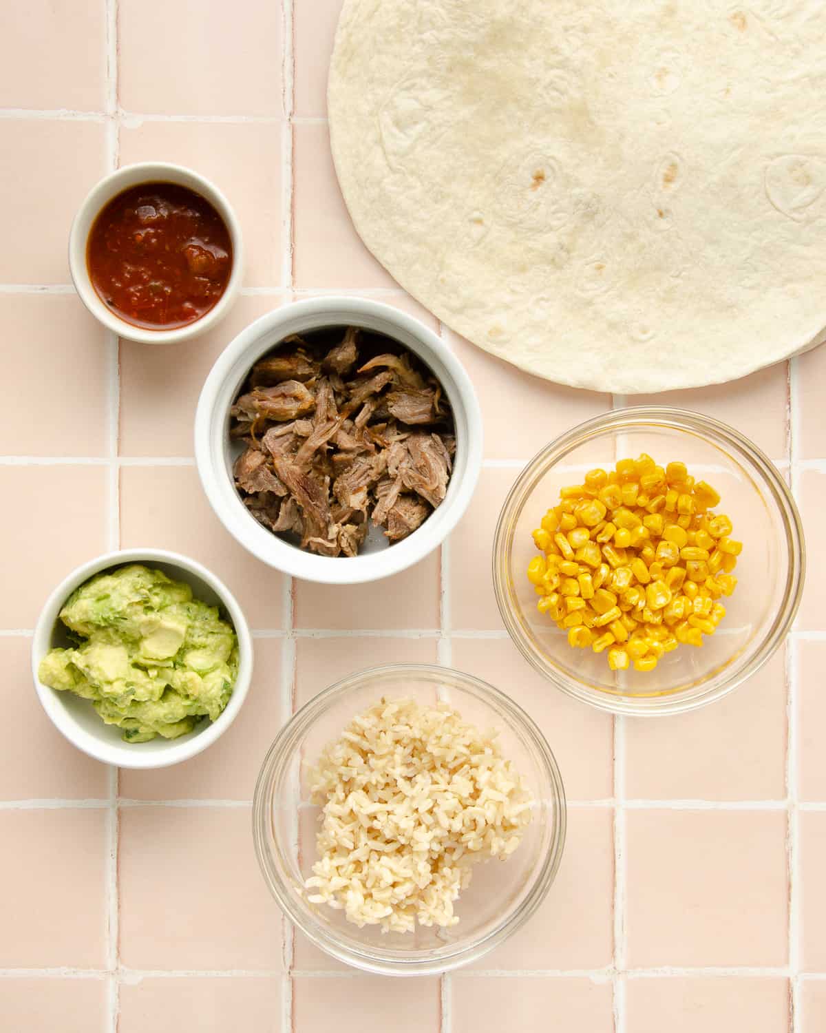 Ingredients needed for carnitas burritos: salsa, pork carnitas, tortilla, corn, guacamole, and rice in glass containers on a pink tile countertop. 
