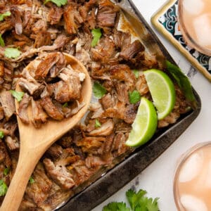 A baking sheet with crispy carnitas, garnished with lime wedges.