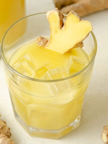 A glass of ginger juice with ice and sliced fresh ginger. More ginger juice and fresh ginger in the background.