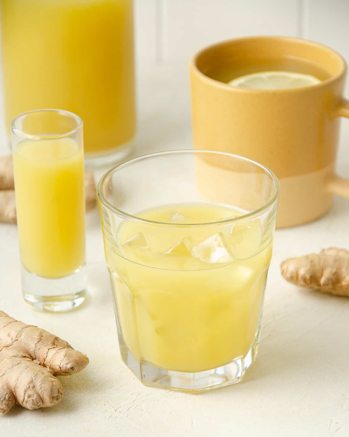 A glass of fresh ginger juice with ice, next to a shot of ginger juice and a mug of ginger tea.