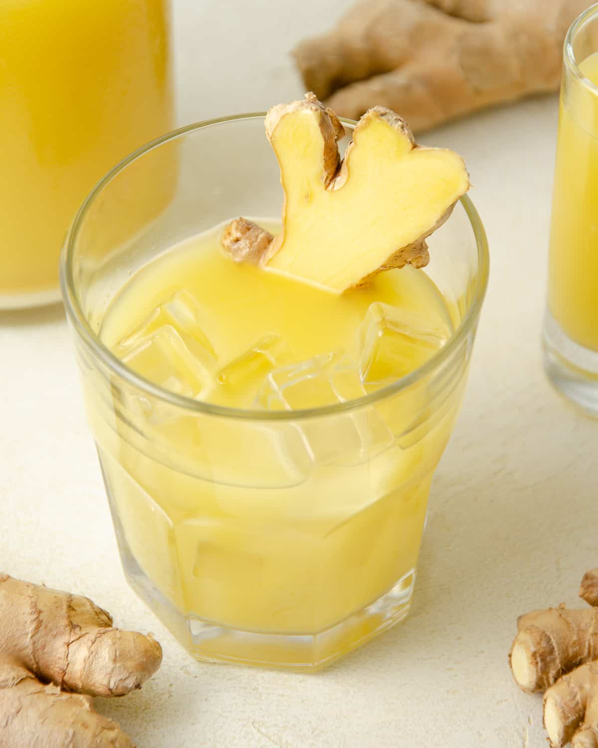 A glass of ginger juice with ice and sliced fresh ginger. More ginger juice and fresh ginger in the background.