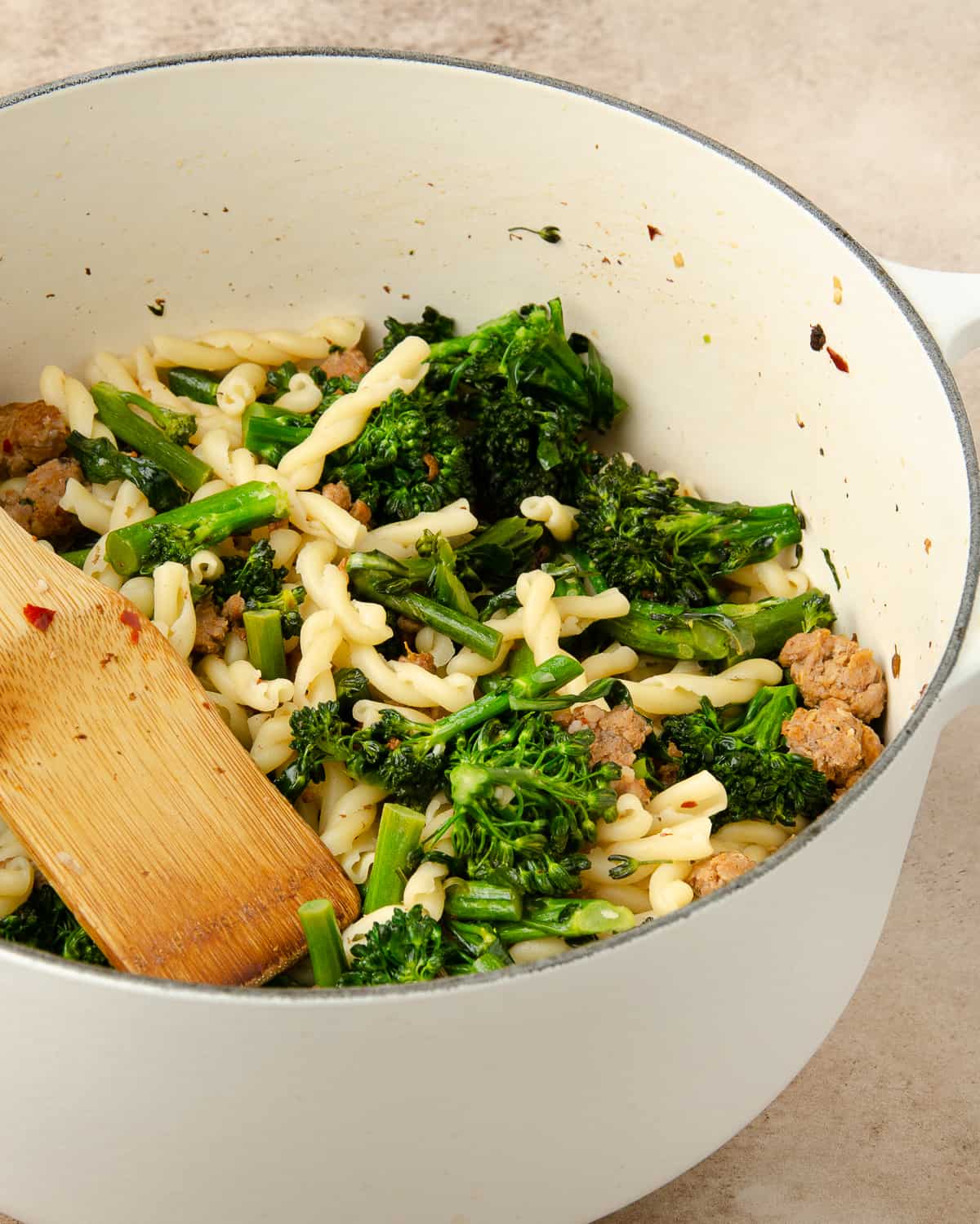 A wooden spoon stirring a white pot of sausage and broccolini pasta. 