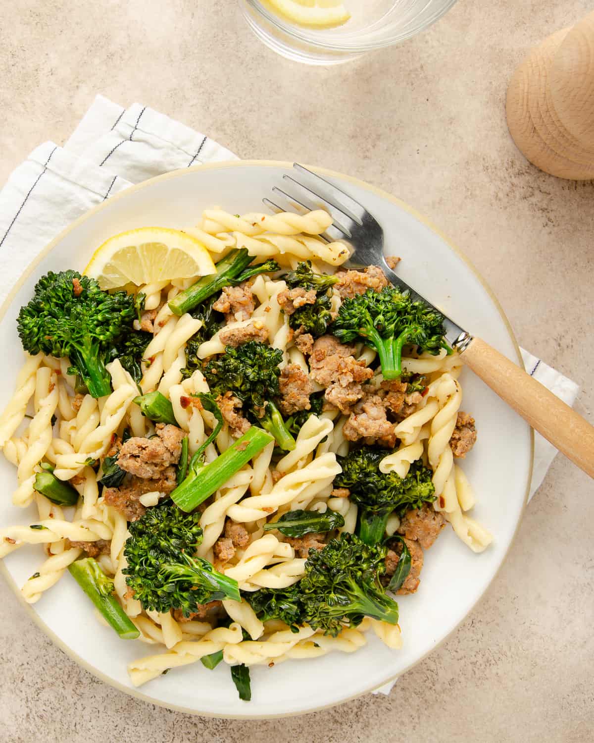 A white bowl full of sausage and broccolini pasta with a lemon wedge, and fork. 