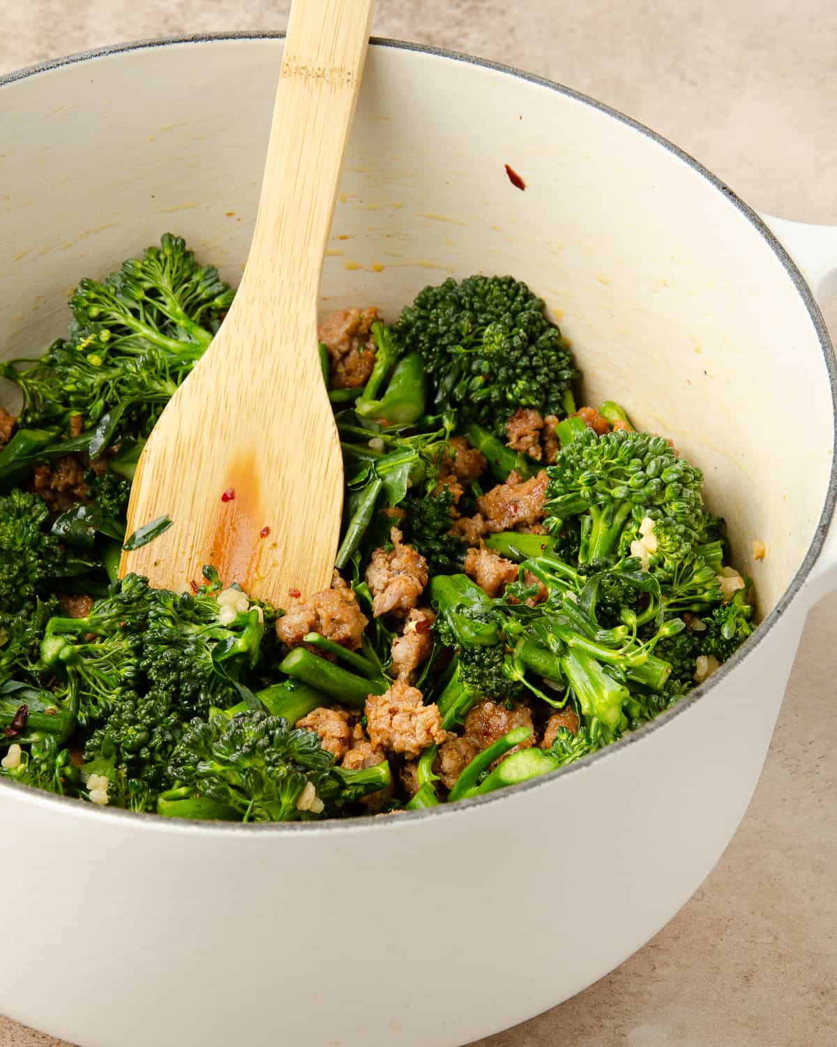 A wooden spoon stirring a white pot of sausage and broccolini pasta. 