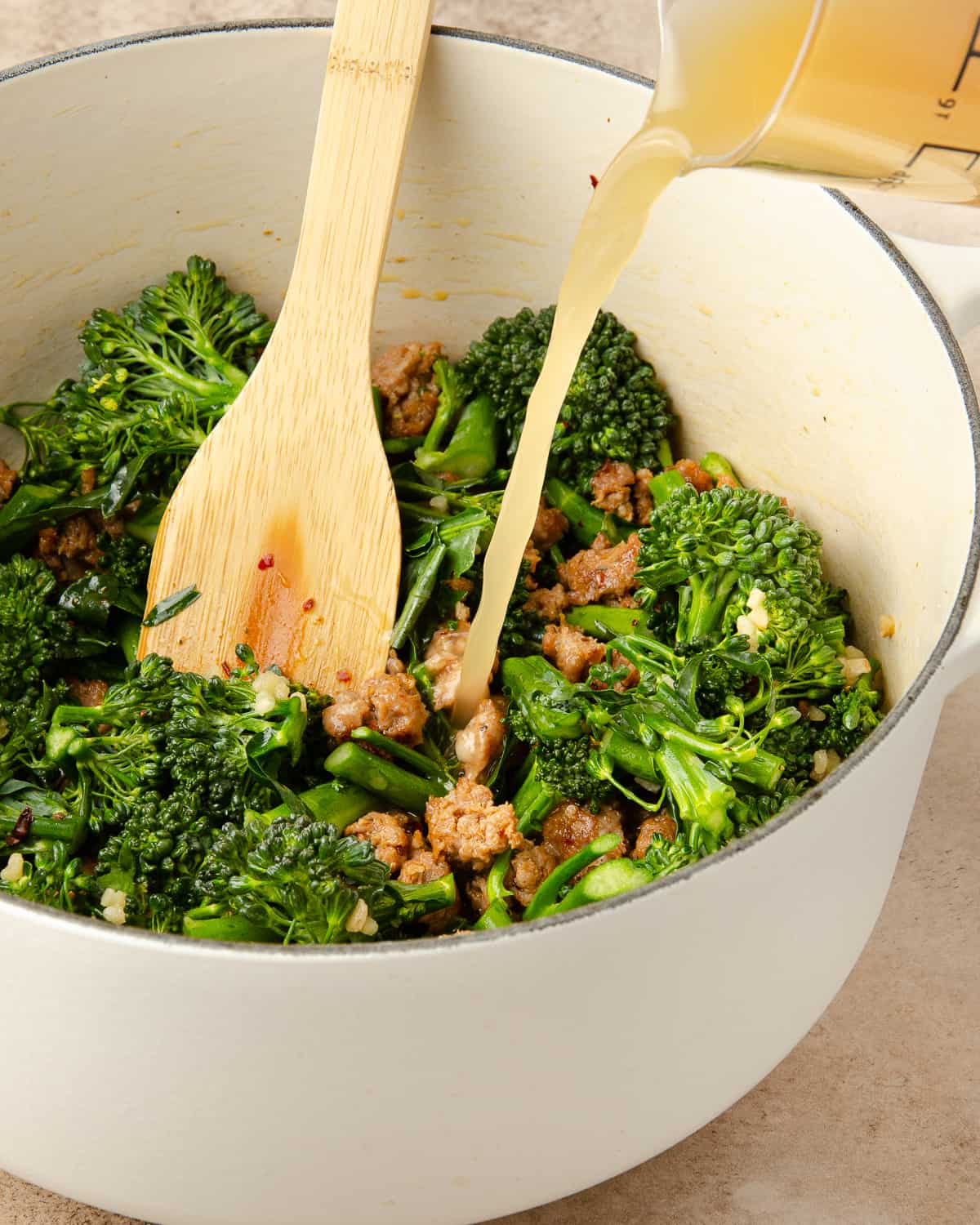 Pouring in broth to a pot of cooked sausage and broccolini. 