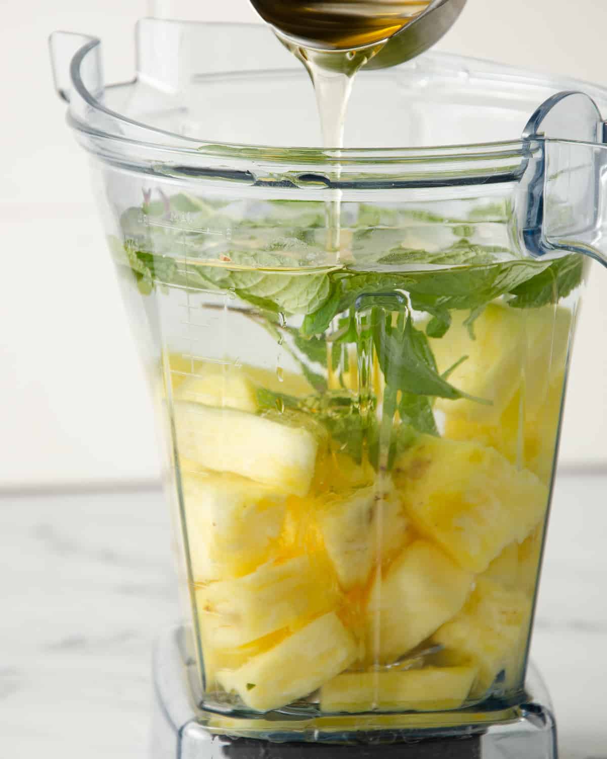 Pouring agave nectar into a blender filled with pineapple, water and mint.