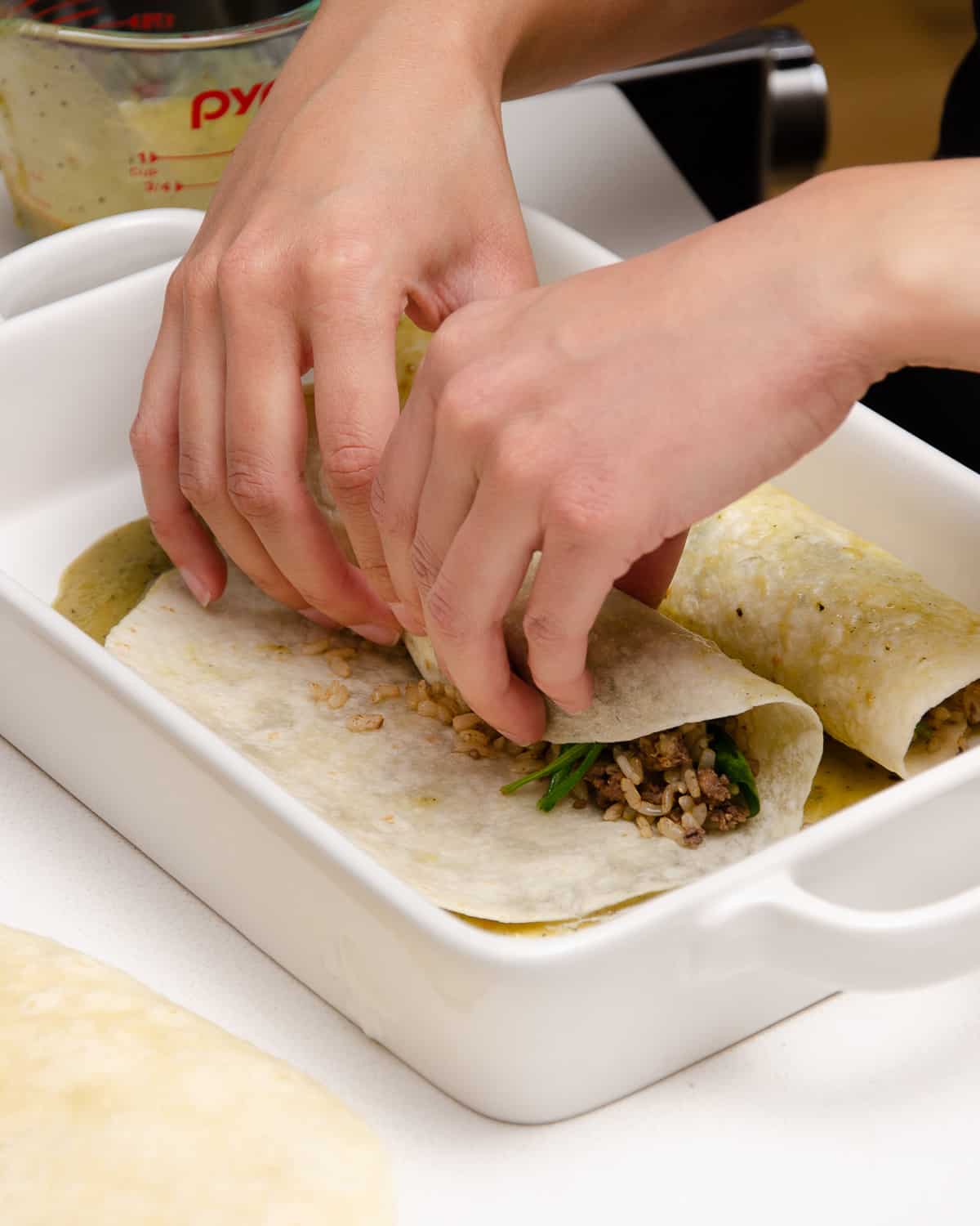 Rolling up beef enchiladas in a white casserole dish.