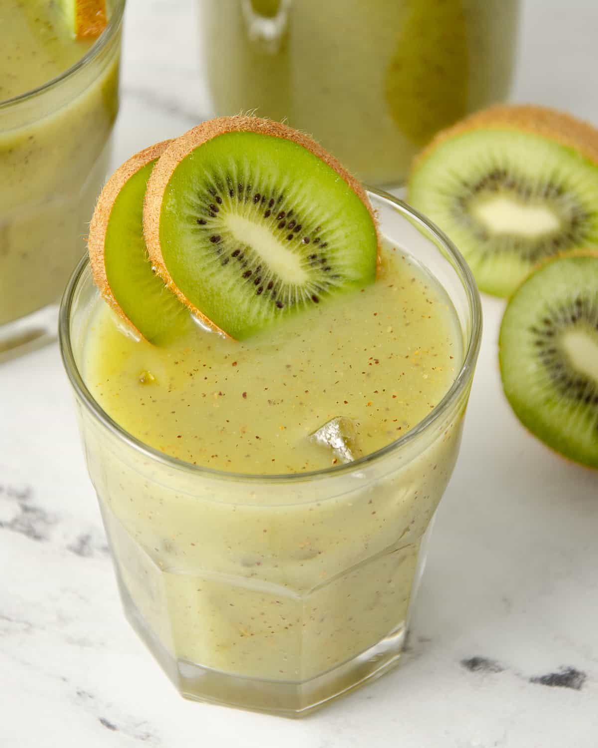 A glass of fresh kiwi juice with kiwi slices, and a pitcher of kiwi juice in the background.