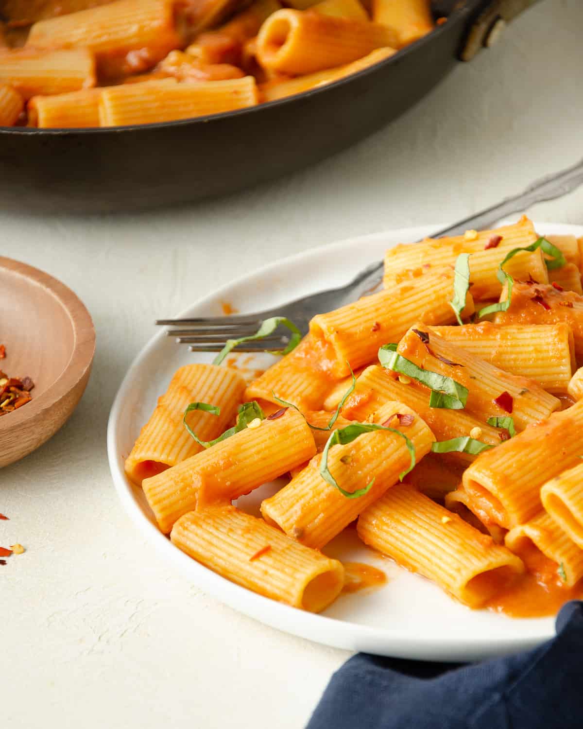 A white plate with rigatoni and dairy free pink pasta sauce, topped with basil and red pepper flakes. A skillet in the background.