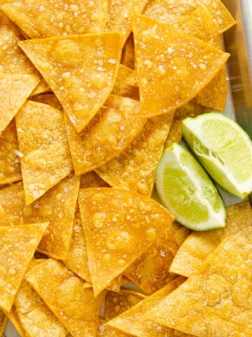 A close up of salted gluten free tortilla chips on a baking sheet with lime pieces.