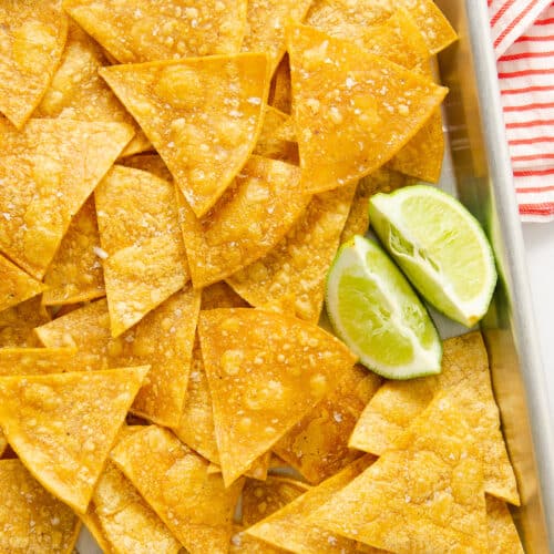 A close up of salted gluten free tortilla chips on a baking sheet with lime pieces.