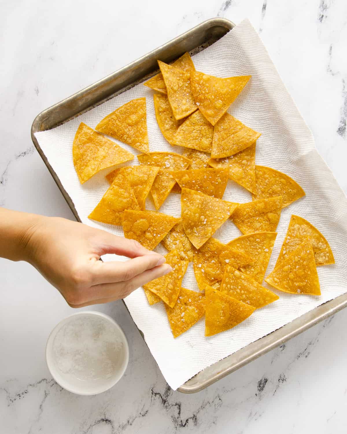 A hand pouring salt to season baked tortilla chips on a paper towel lined baking tray.