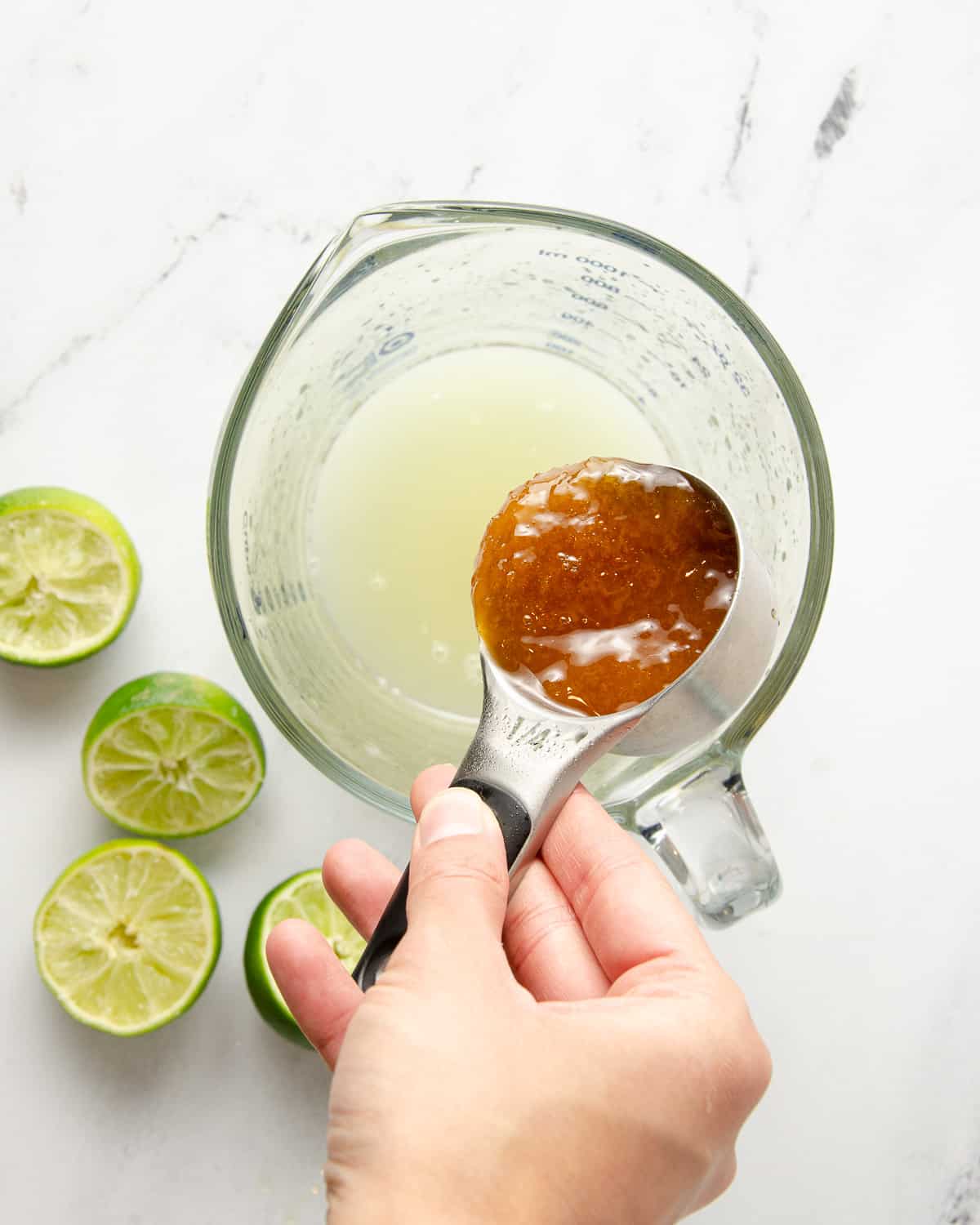A hand pouring honey into a measuring cup filled with lime juice.