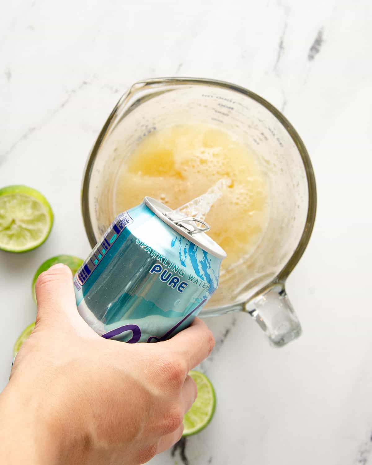 A hand pouring a can of sparkling water into a large glass measuring cup.