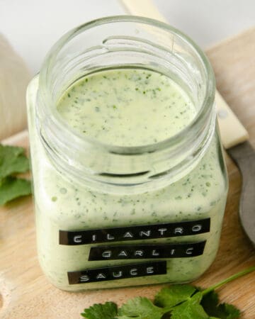 Blended cilantro garlic sauce in a jar on a cutting board with cilantro leaves next to it.