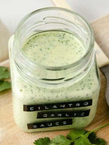 Blended cilantro garlic sauce in a jar on a cutting board with cilantro leaves next to it.