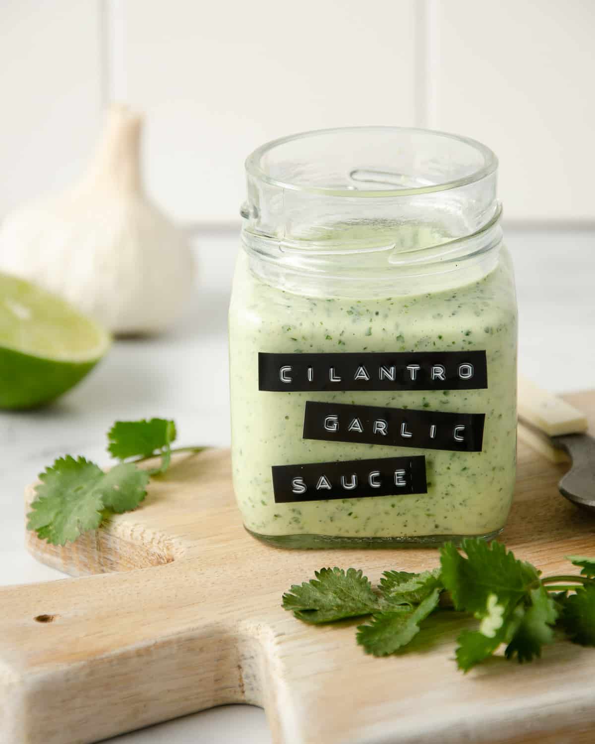 Blended cilantro garlic sauce in a jar on a cutting board with cilantro leaves next to it and limes and garlic in the background.