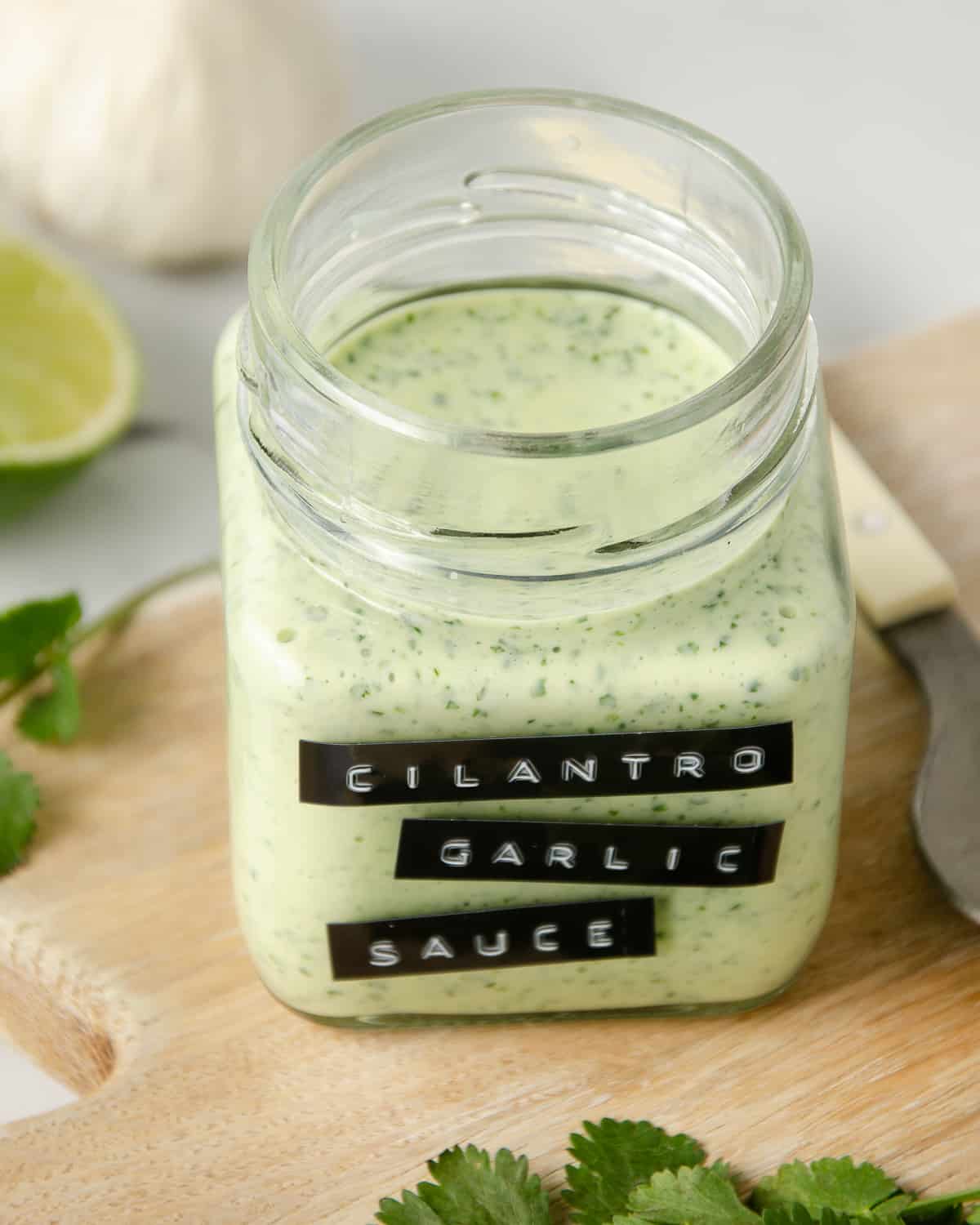 Blended cilantro garlic sauce in a jar on a cutting board with cilantro leaves next to it and limes and garlic in the background.