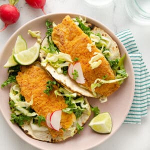Crispy fish tacos on a plate topped with lime, slaw, and radishes.
