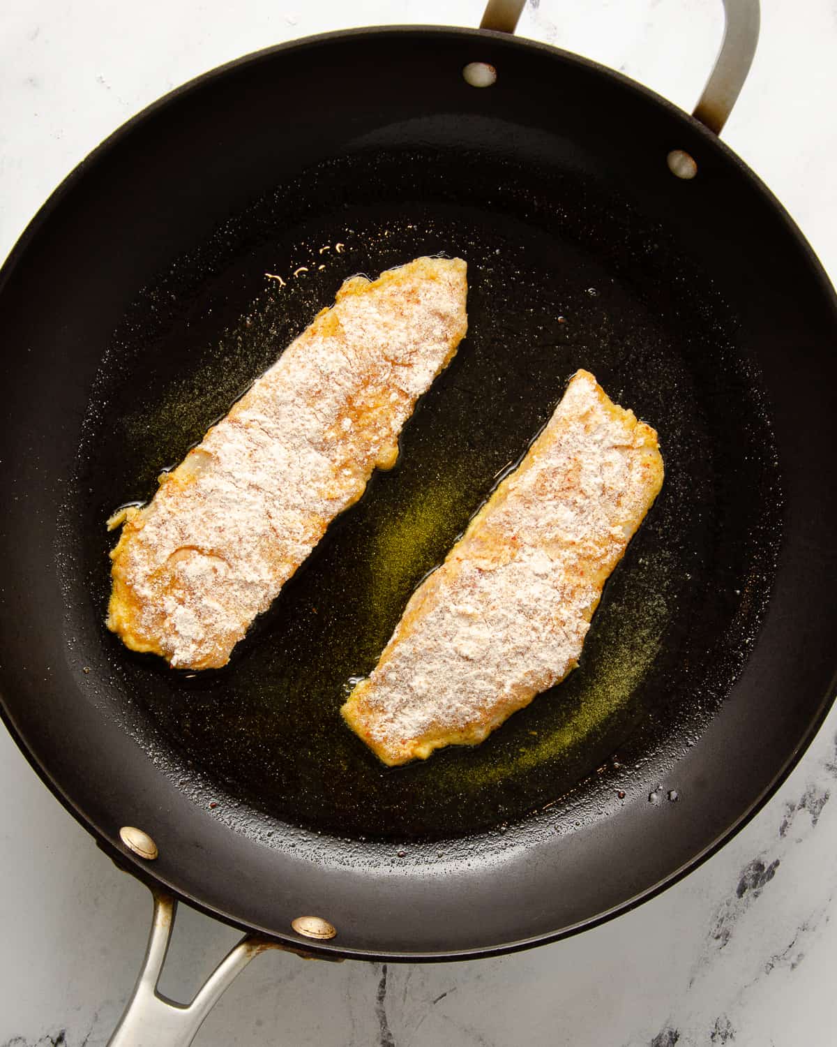 Two pieces of tilapia frying in a frying pan.