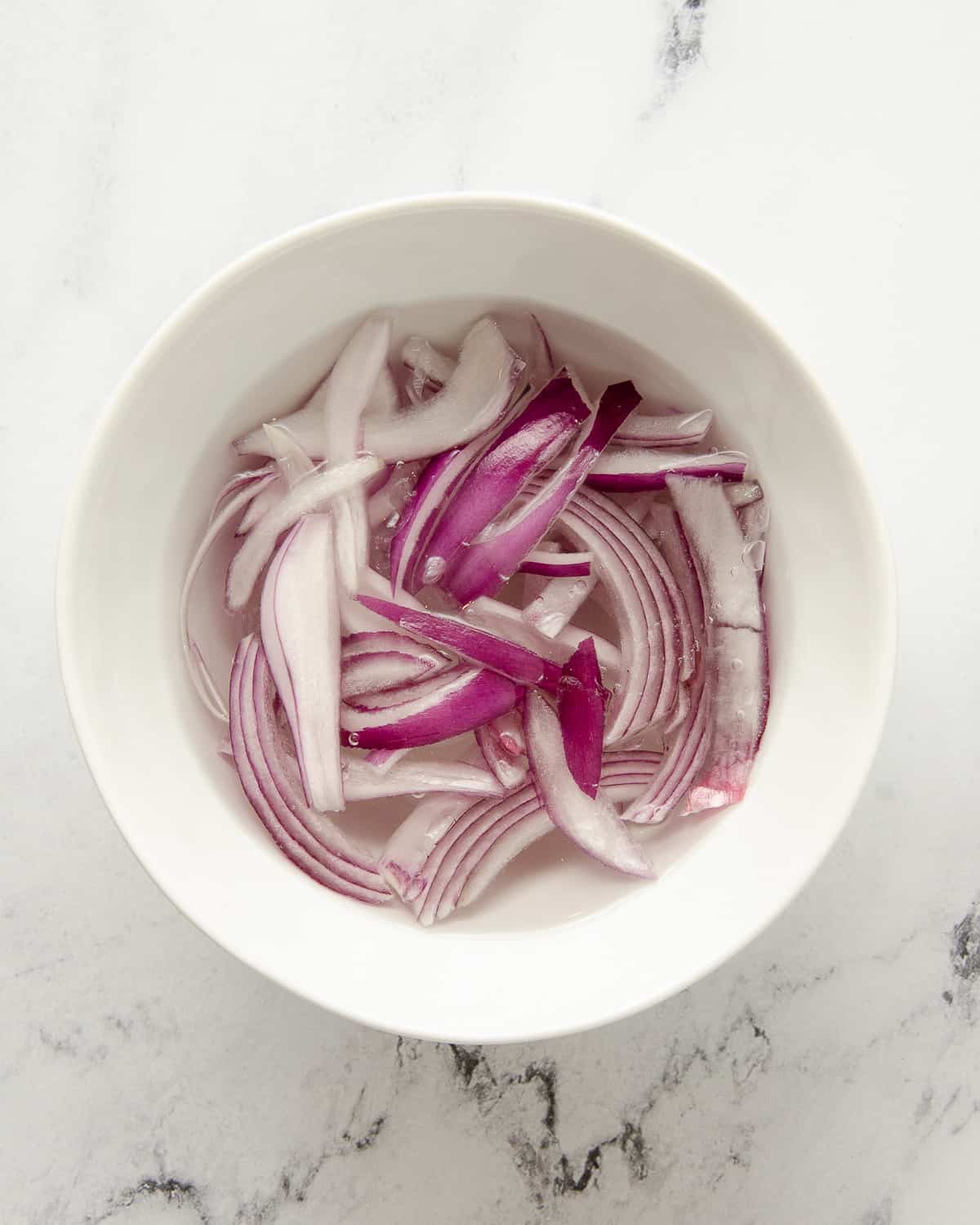 Thin sliced red onion in a bowl submerged in water to reduce the harshness of the onion.