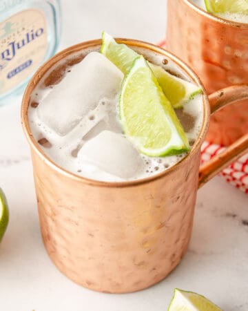 A featured image of a tequila mule in a copper mug with two lime wedges inside.