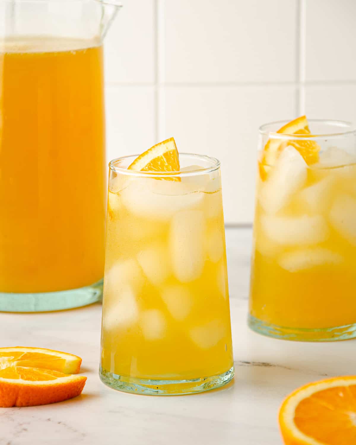 Two tall glasses of orange agua fresca with an orange wedge garnish and pitcher to the side.