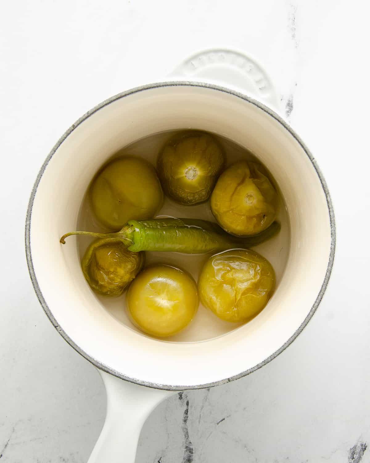 Boiled tomatillos and a serrano pepper in water in a sauce pot.