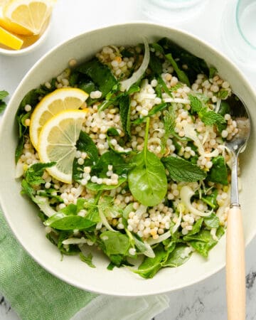 A bowl full of couscous salad topped with spinach, fresh mint, and lemon slices with a serving spoon.