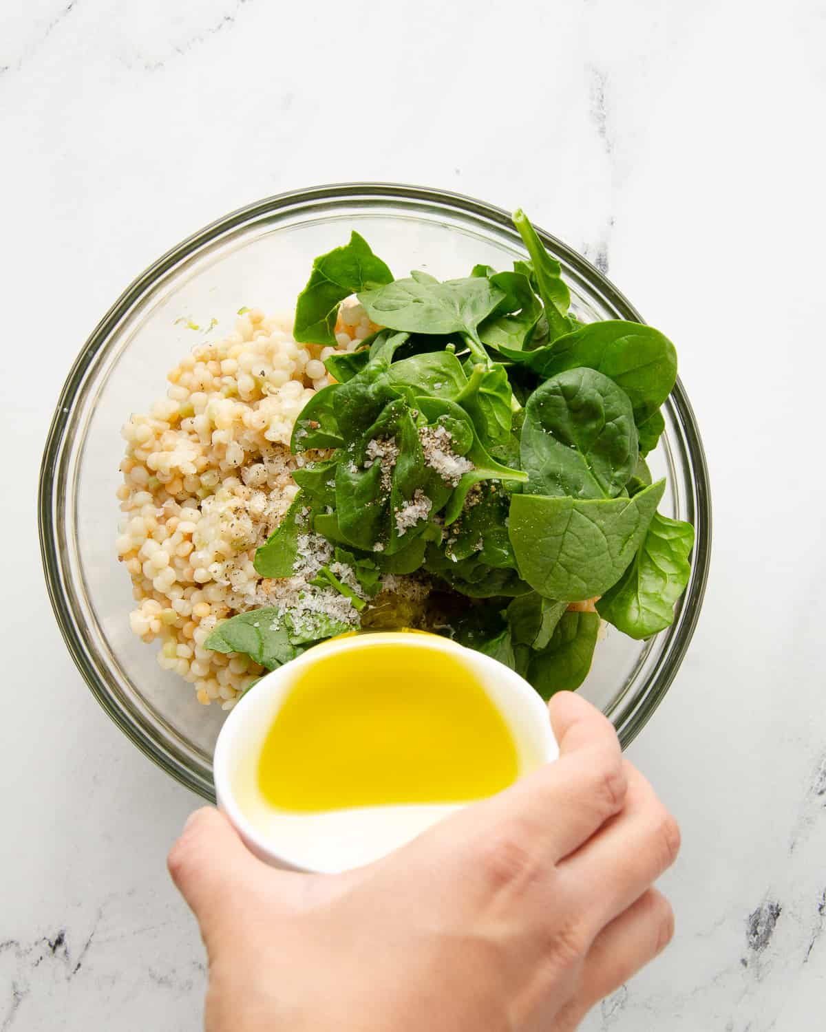 A hand pouring olive oil into a glass bowl with couscous and spinach.