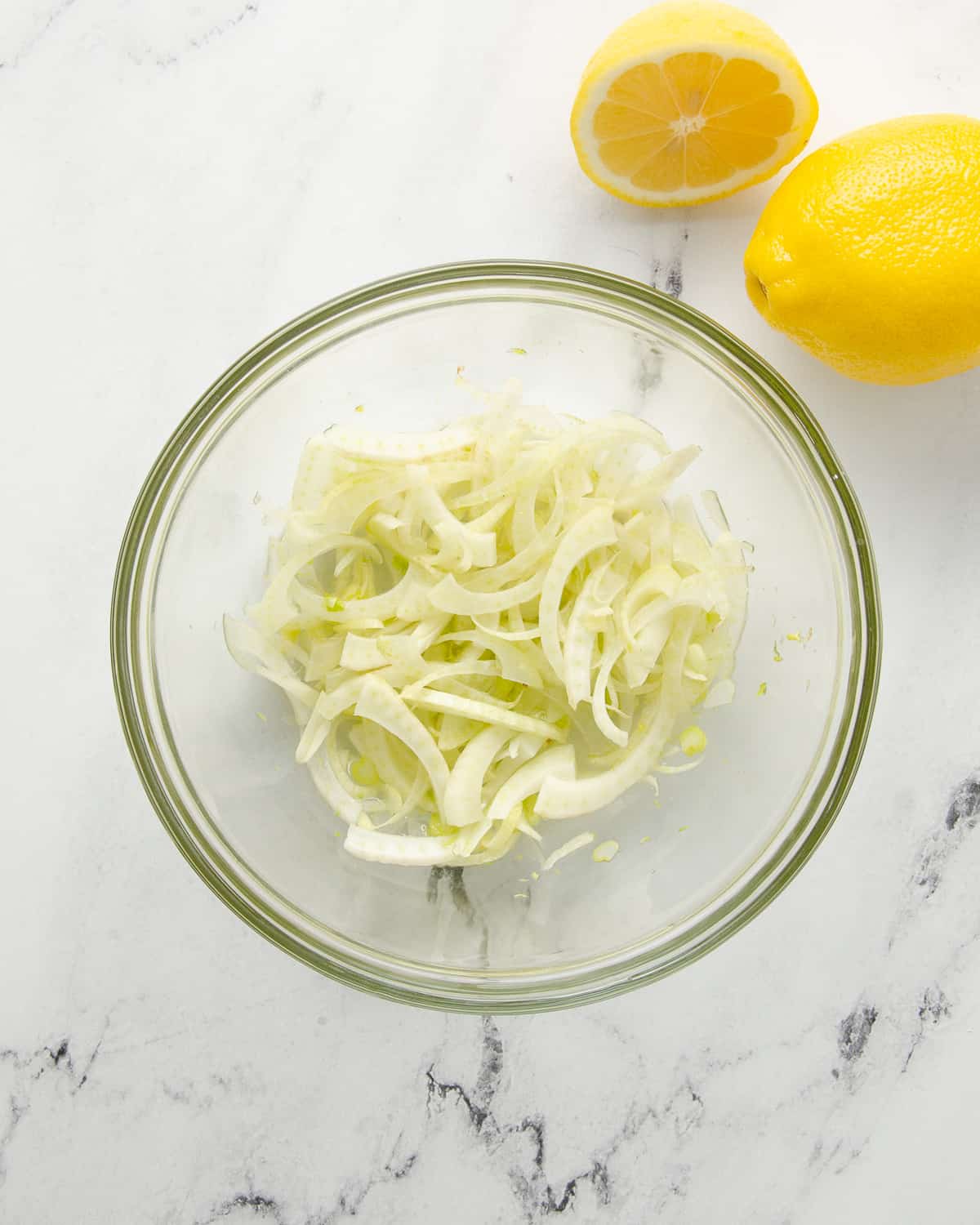 A glass bowl with shaved fennel next to a lemon and a lemon wedge.