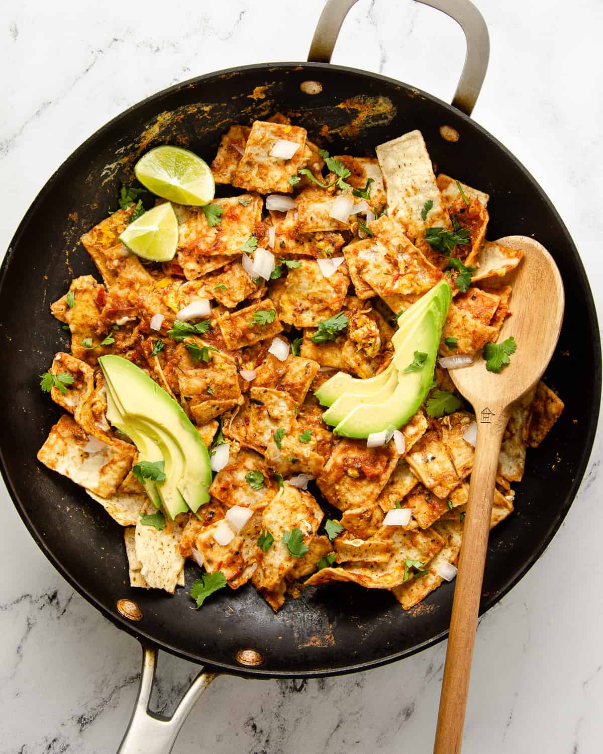 A skillet in view with finished chilaquiles topped with avocado slices, diced onion and cilantro.