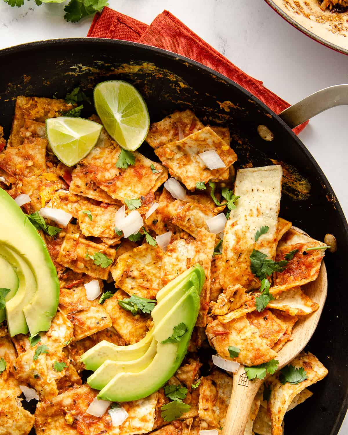 A close up view of chilaquiles rojos in a skillet with a wooden spoon scooping a bite out.