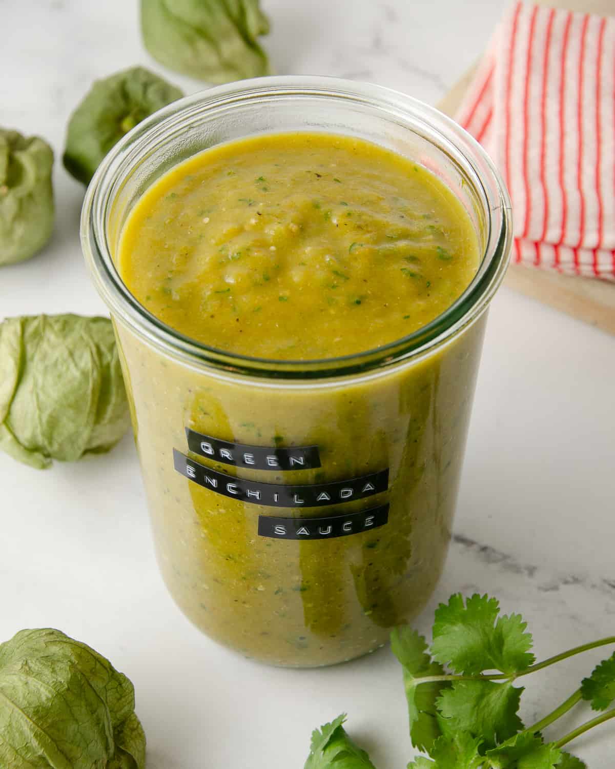 An overhead view of a large jar of green enchilada sauce with tomatillos and cilantro to the side.