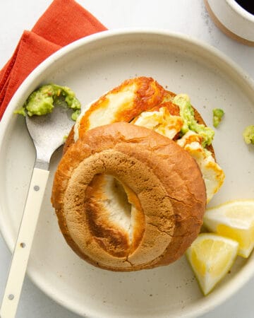 A bagel with halloumi and avocado on a white plate with a red napkin.