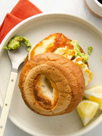 A bagel with halloumi and avocado on a white plate with a red napkin.