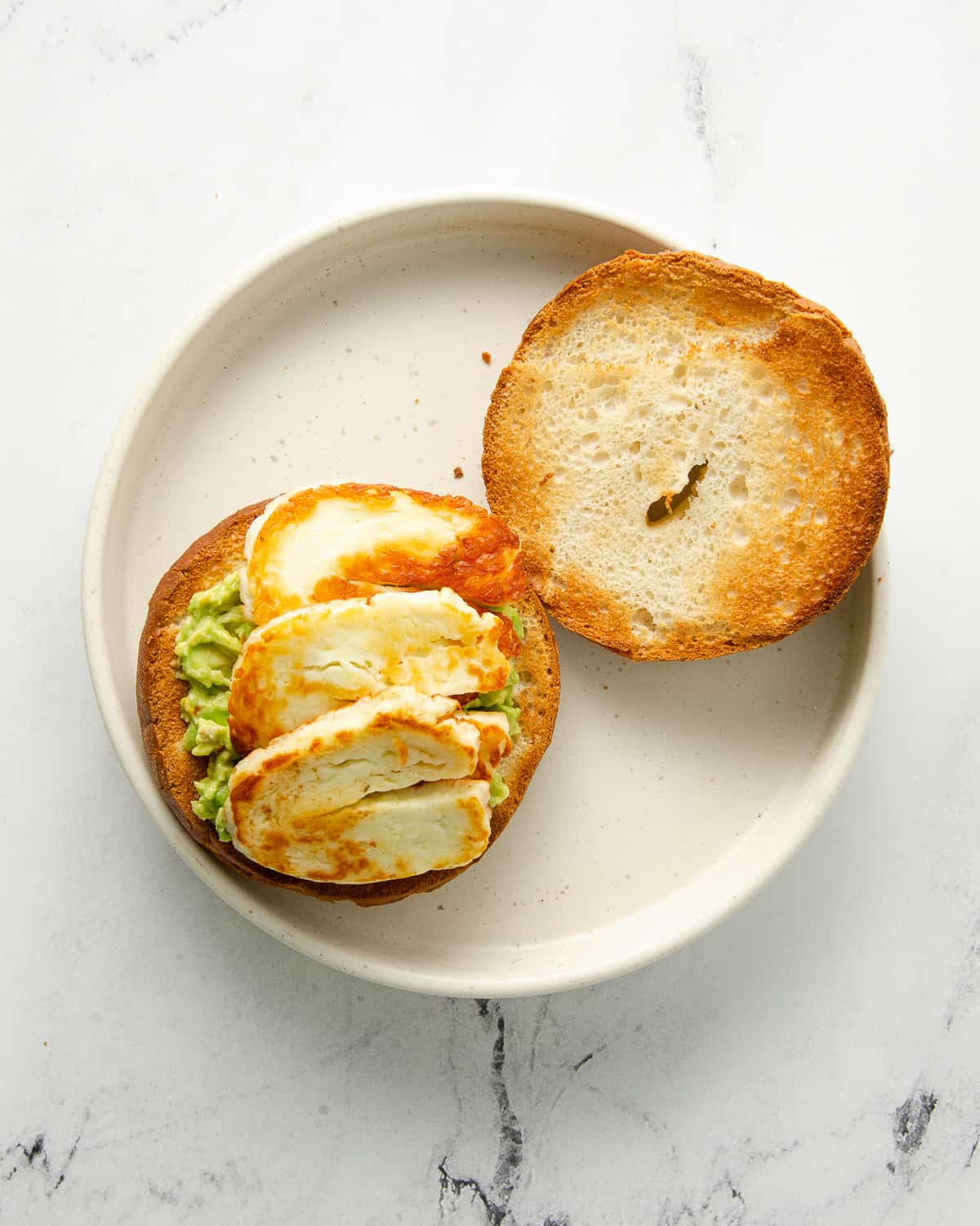 Two halves of a toasted bagel with mashed avocado and halloumi on one side on a white plate.