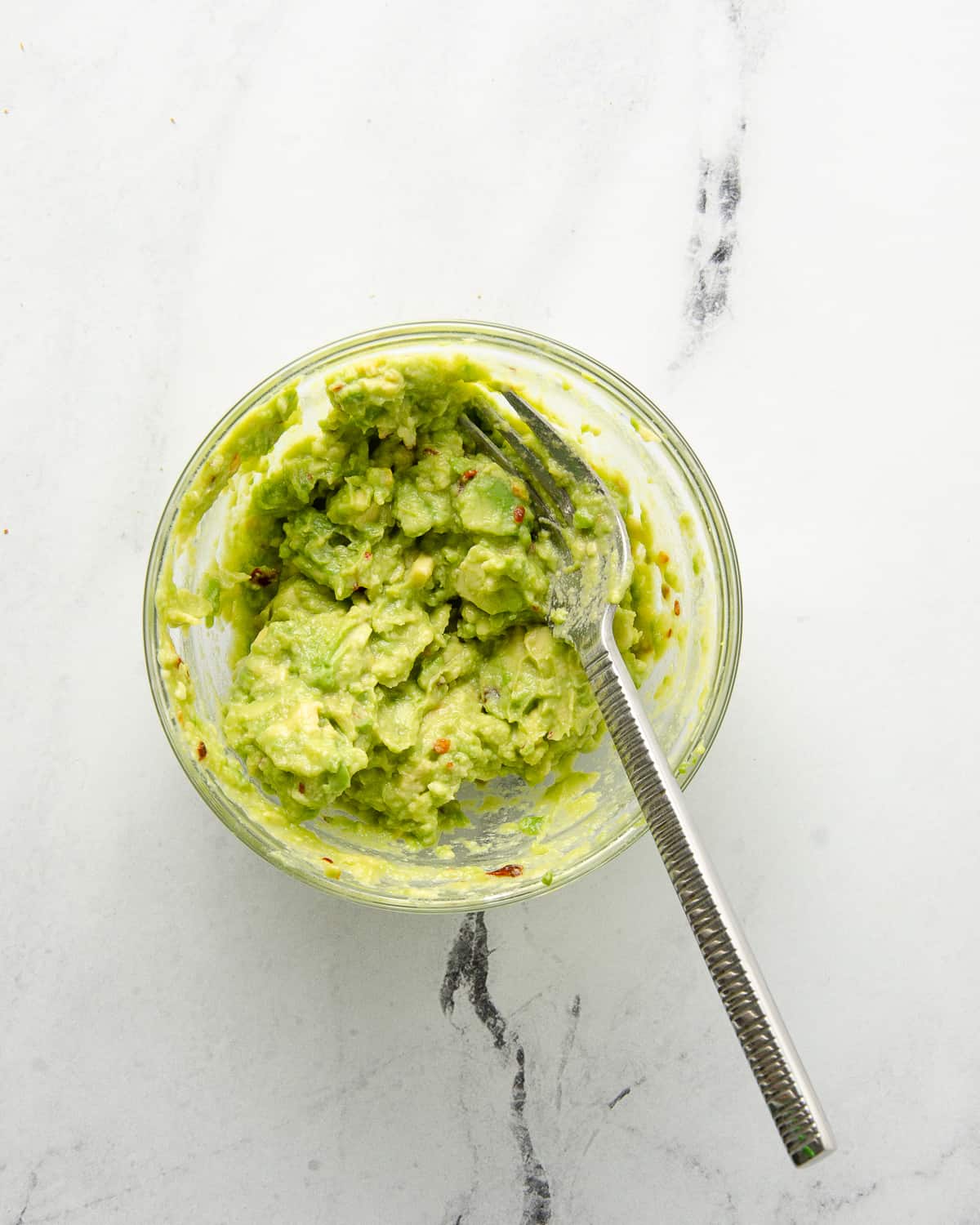 A glass bowl with mashed avocado and a fork.