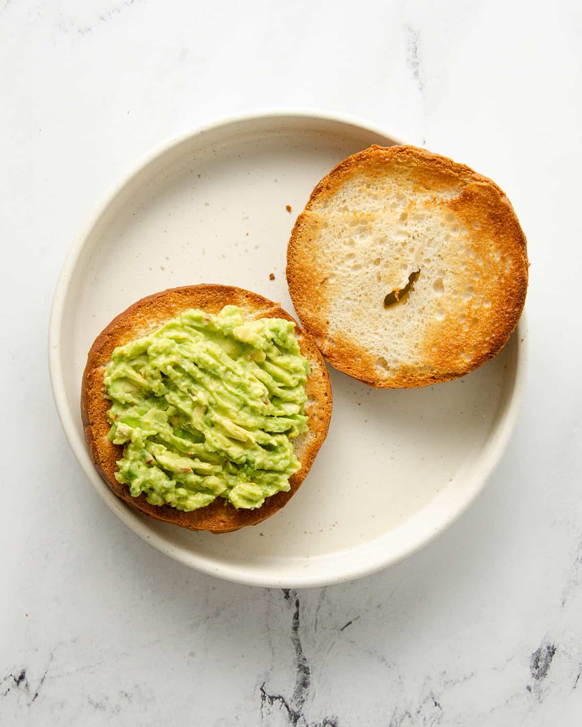 Two halves of a toasted bagel with mashed avocado on one side on a white plate.