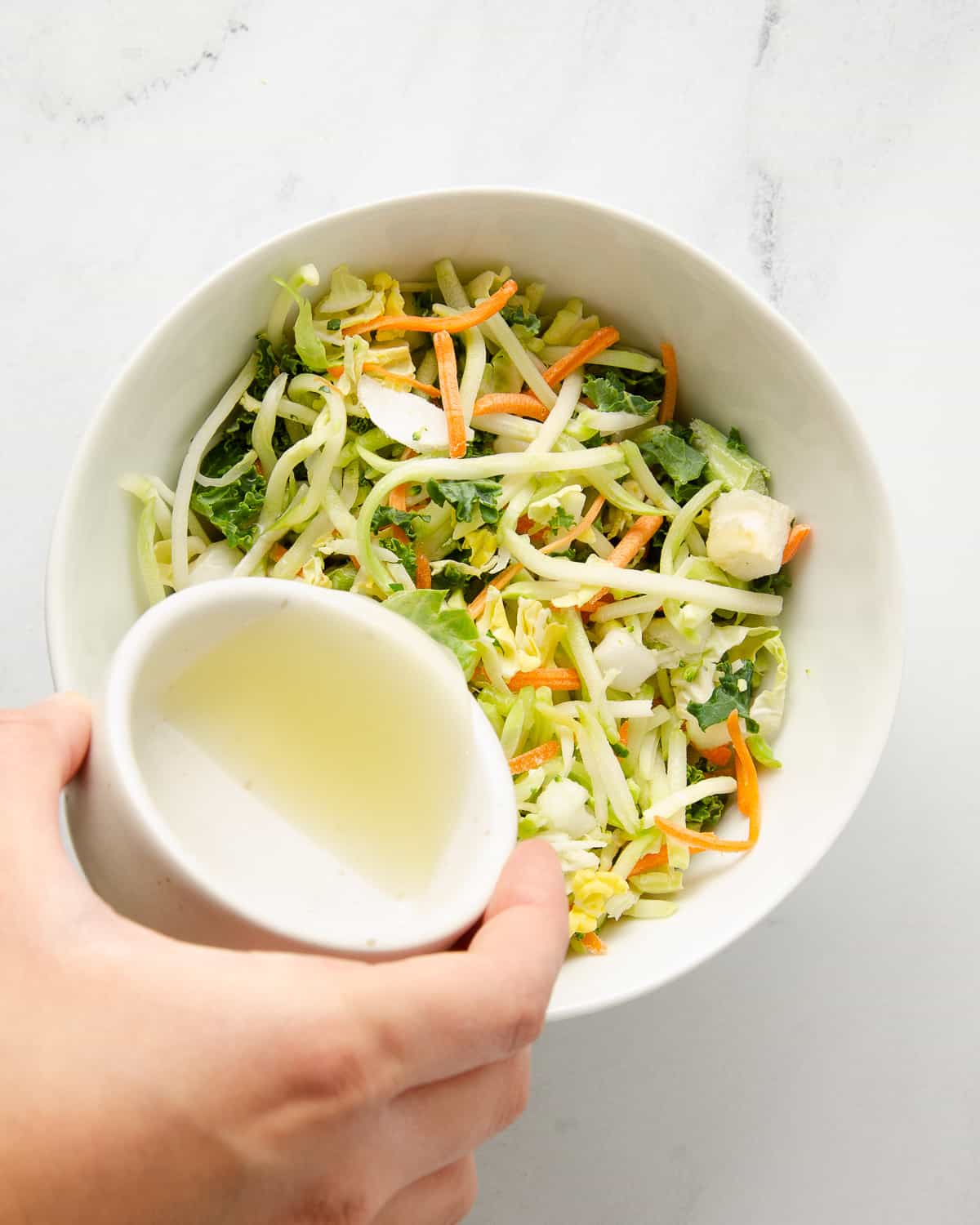 A hand pouring lime juice onto slaw mix in a white bowl.