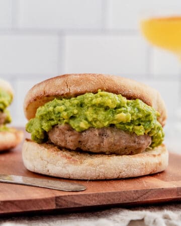 Breakfast burger on an english muffin topped with guacamole on a cutting board.