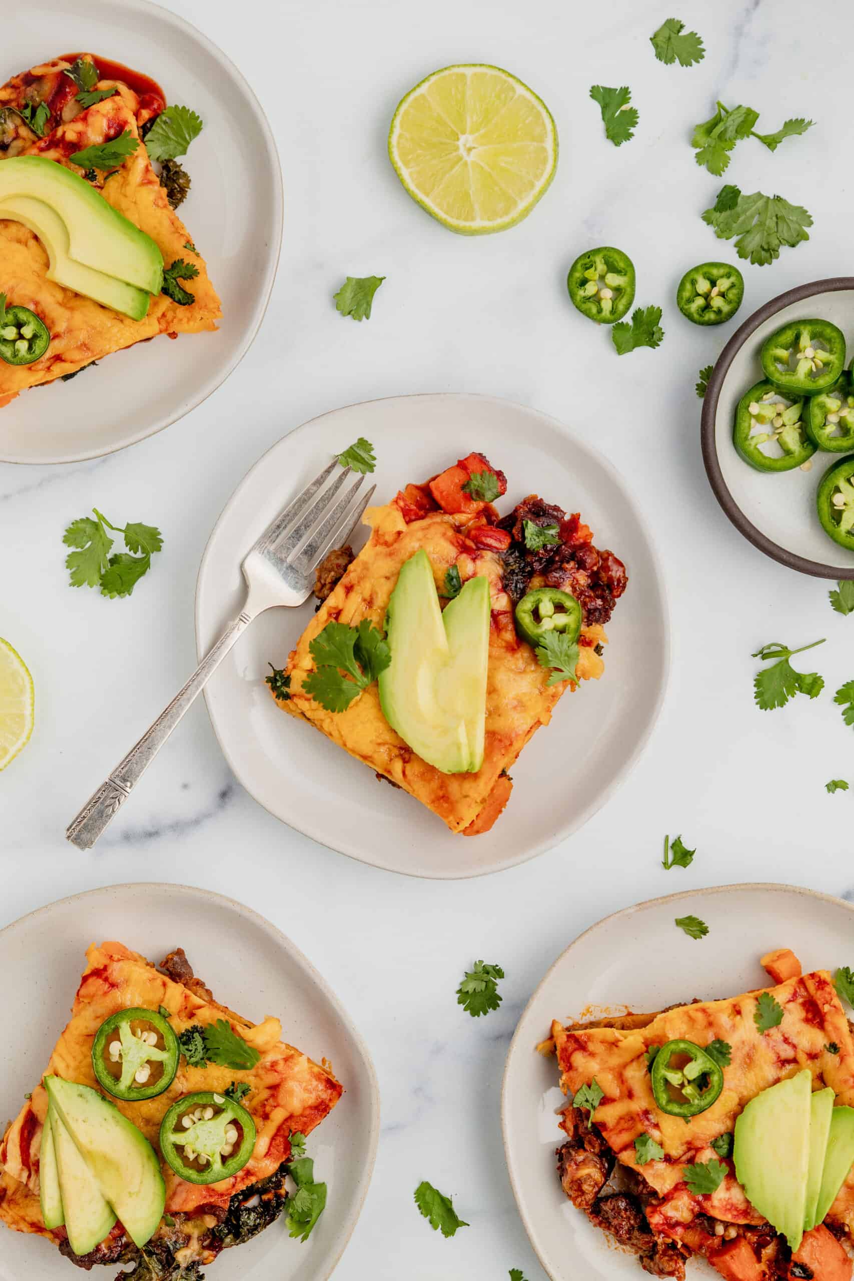 Four plates with slices of gluten free enchilada casserole topped with cilantro, limes, and avocado.