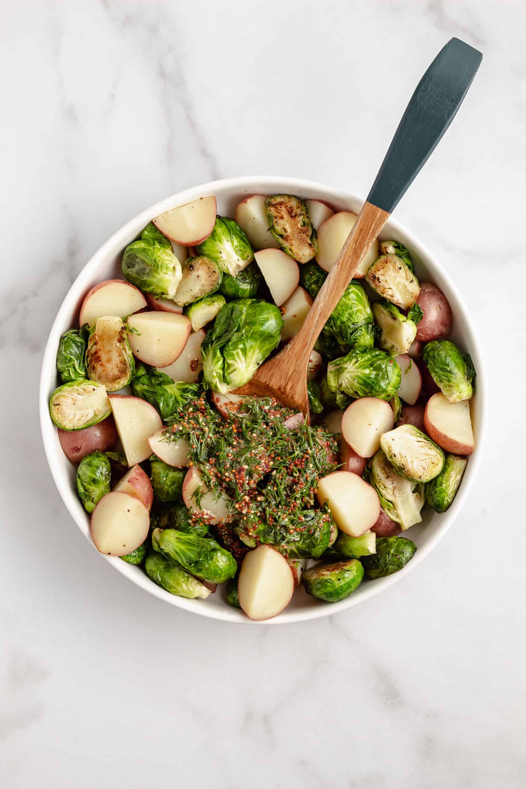 A white bowl with potatoes and Brussels sprouts topped with mustard dill dressing with a serving spoon.