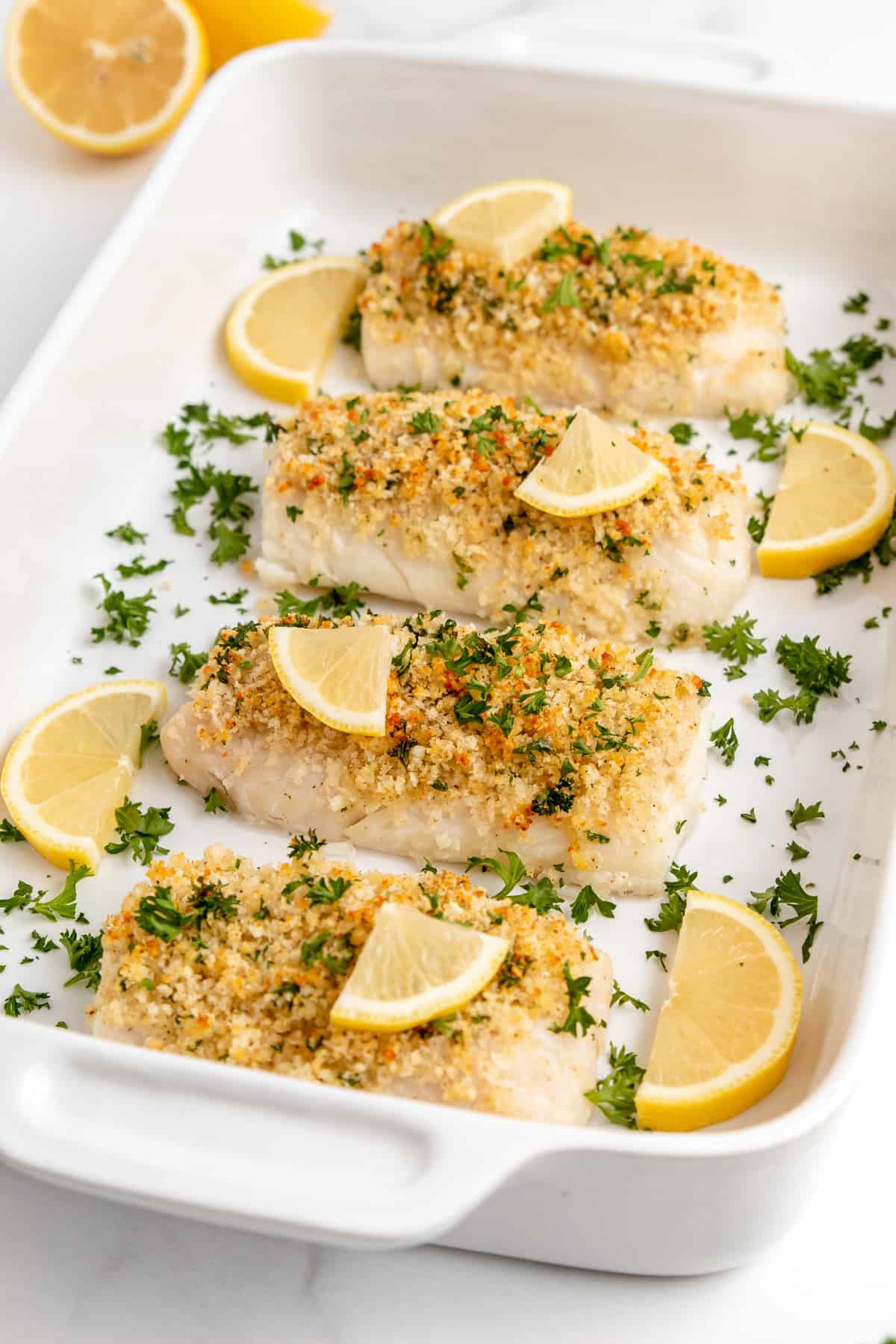 Up close view of panko coated baked cod fillets with lemon and parsley in a baking dish.