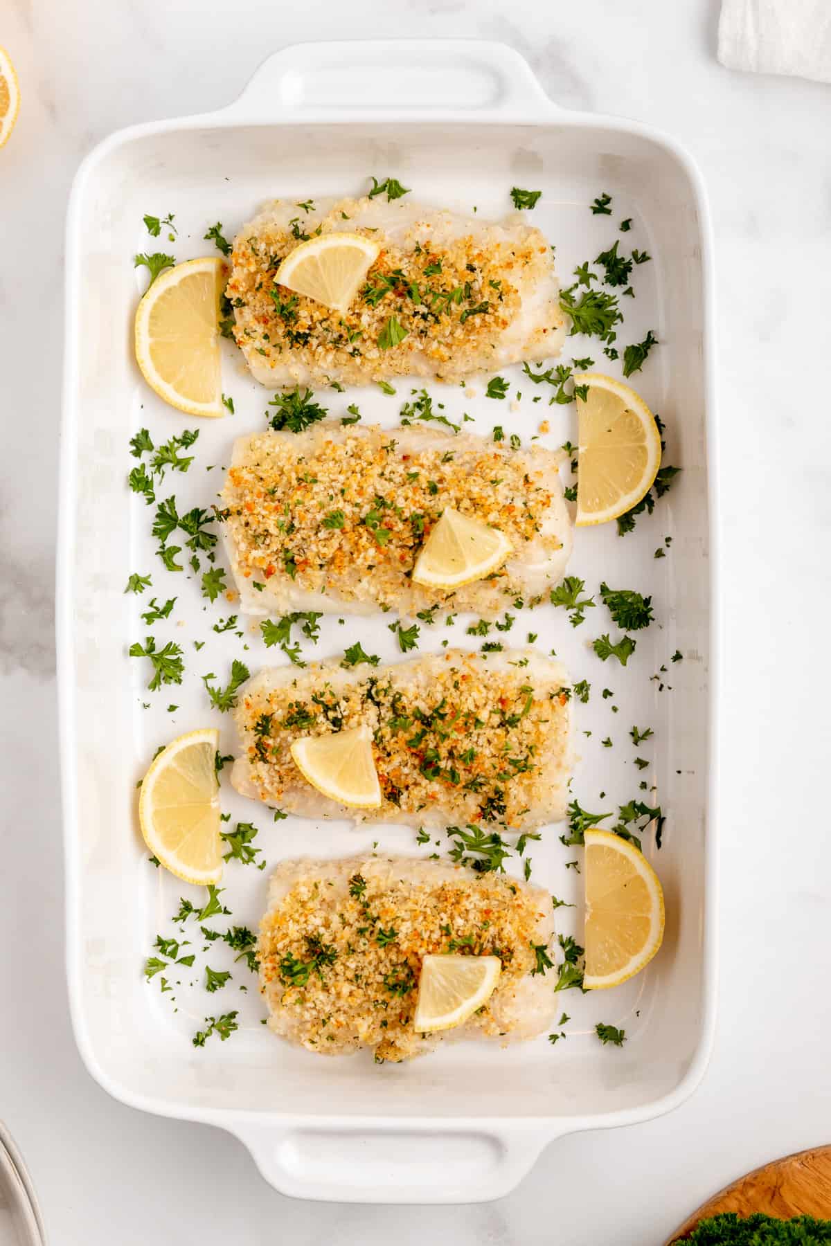 A baking dish with pieces of panko coated baked cod fillets topped with lemon and parsley.